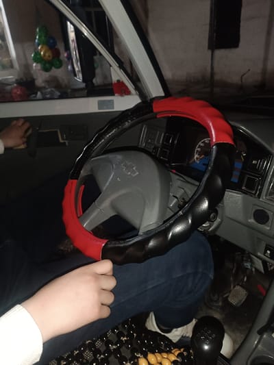 The image captures a close-up view of the interior of a car, primarily focusing on the driver's area, including the steering wheel, dashboard, and the driver's hands and legs.
In the foreground and center, a gray steering wheel is visible, covered with a two-tone steering wheel cover that is black on the sides and red on the top and bottom sections. The cover has a textured, slightly bumpy surface. A faint Chevrolet logo is visible on the center of the gray steering wheel itself, beneath the cover.
A person is seated in the driver's seat, with their lower body and hands visible. They are wearing blue jeans and white shoes. One hand, wearing a white long-sleeved top, rests on the left side of the steering wheel cover, while the other hand is seen further to the left, near the car's door panel, possibly resting on the armrest or door handle. A black and yellowish-orange beaded seat cover is partially visible on the seat beneath the person's legs.
To the right of the steering wheel, a basic dashboard and instrument cluster are visible. A speedometer or tachometer gauge has a prominent blue background on its lower portion. Other dark-colored buttons and switches are arrayed across the dashboard. Below the dashboard, the car's foot pedals (accelerator, brake, and clutch) are visible on the floor. To the right and slightly below the driver's right knee, a black manual gear shift knob with visible gear numbers (e.g., "1", "2", "3", "4", "5", "R") is present.
Through the front windshield, a dimly lit background, possibly an indoor garage or a wall, can be seen. An interior rearview mirror is visible, reflecting parts of the car's interior or what's outside the side window. A multi-colored ornament, consisting of several small, spherical objects (green, red, yellow, blue), hangs from the rearview mirror or near the windshield.
The overall lighting is dim and appears to be artificial, suggesting the image was taken indoors or during nighttime. The setting indicates the car is likely stationary.