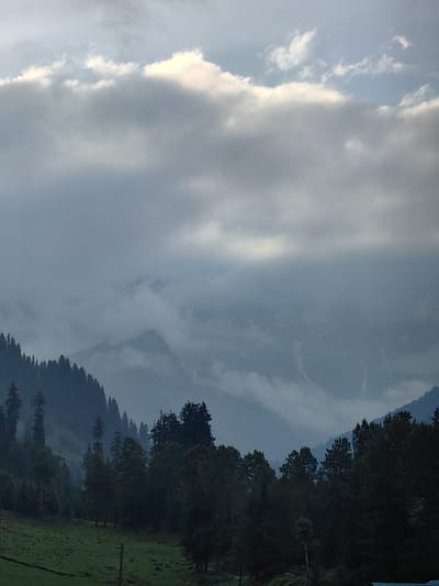The image depicts a panoramic view of the Solang Valley in India, characterized by a dramatic interplay of light and shadow. Towering, mist-shrouded mountains dominate the background, their peaks partially obscured by thick, ethereal clouds. These clouds, a mix of dark gray and luminous white, create a sense of depth and atmospheric drama, with hints of sunlight breaking through in the upper portion of the sky.
In the middle ground, dense forests of evergreen trees, primarily pine and deodar, blanket the steep slopes of the mountains. The trees are dark and imposing, their silhouettes sharp against the lighter hues of the sky and distant mist. The forest canopy appears lush and untouched, suggesting a remote and wild environment.
The foreground features a gently sloping meadow of vibrant green grass, marked by subtle undulations and a few scattered rocks. A dark, winding path or track is visible across the meadow. A few utility poles with wires are present, suggesting the presence of some human infrastructure, though no people are visible in the frame. The overall impression is one of serene, majestic natural beauty, with a slightly melancholic or mysterious atmosphere due to the overcast sky and misty mountains.