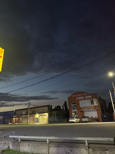 This media file depicts a street scene at dusk in Yegorlykskaya, Russia. The image is oriented vertically, with the sky taking up the left half and the street and buildings on the right.

The primary subjects are two buildings situated along the roadside. The building on the left is a brick structure with a prominent sign that reads "АПТЕКА" (Pharmacy) and "НАДЕЖНАЯ ПОМОЩЬ" (Reliable Help). It features large windows and a roll-up security shutter. Two cars, a white hatchback and a silver hatchback, are parked in front of this building. The building on the right appears to be a commercial establishment with a brightly lit facade displaying various advertisements and displays. A bus stop sign is visible in front of this building.

The scene is illuminated by a street light overhead and the interior lighting of the businesses. The sky is a deep blue, transitioning to lighter shades, with scattered clouds and what appear to be power lines crisscrossing the frame. The overall atmosphere suggests a quiet evening in a small town. The time of day is clearly dusk, as indicated by the dark sky and artificial lighting.

Visible text includes "АПТЕКА," "НАДЕЖНАЯ ПОМОЩЬ," and other Cyrillic lettering on the signs of the businesses, though some are partially obscured or difficult to read. A yellow sign with a red symbol is partially visible at the bottom of the frame, possibly indicating a pedestrian crossing.