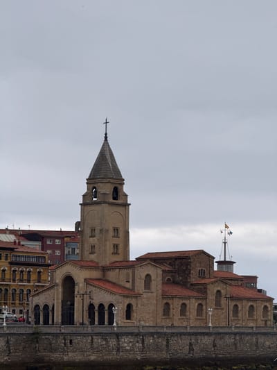 A vertical, eye-level shot depicts a stone building with a steeple and multiple levels in Gijón/Xixón, Spain. The sky is overcast and mostly fills the left side of the frame.

The building's steeple, topped with a cross, extends into the sky. The structure has a symmetrical design with arched windows and is constructed of light-colored stone. Red tile roofs cover various sections of the building. Behind the steeple is a light-colored building that is multiple stories tall with windows.

On the right side of the frame, there is a stone wall with some moss growing on it. Further up the building, a weathervane with flags extends from the top of the building. The lower floors have arched entryways and windows.

At the bottom of the frame, other buildings and some street-level details are visible, including some cars and building facades.