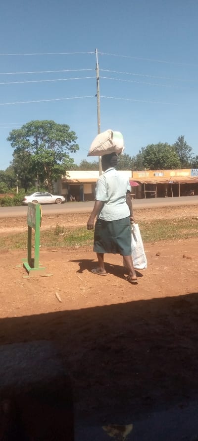 The image shows a woman walking on a dirt road. The woman is wearing a light blue and white checkered shirt, a green skirt, and sandals. She is carrying a white bag and a large bag of goods on her head. The woman is walking towards the right side of the image. In the background, there are trees, a white car, and a structure with a yellow sign. The woman is walking past a green sign with white lettering. The sky is blue with white clouds.  The location is Kivaywa, Kenya, at the coordinates 0.56 latitude and 34.805 longitude.