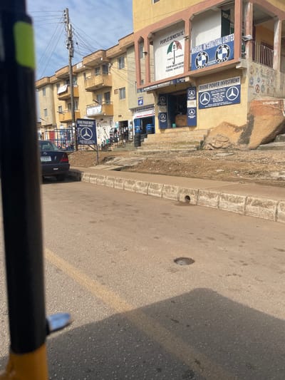 The image captures a street scene in Nigeria, possibly near a stadium given the context. The perspective is from a vehicle, looking towards a two-story building with several shops or offices on the ground floor. The building is painted in shades of yellow and beige.
Several signs are visible on the building. One large blue sign prominently displays the Mercedes-Benz logo and text, including "DIVINE POWER INVESTMENT MERCEDES BENZ" and contact information. Another sign is for "MARSHAL INTERNATIONAL LIMITED" with BMW logos. Further down, a sign reads "PEOPLES DEMOCRATIC" with "DP" logo, indicating a political office, possibly a ward office. Above these shops, windows are visible on the second floor.
The street itself is paved with asphalt, showing some wear and tear, with a concrete curb. A drain grate is visible on the curb. To the left of the building, more of the structure extends upwards, with balconies and windows. A utility pole with numerous transformers and wires is also present, a common sight in Nigerian streetscapes. A dark blue Mercedes-Benz car is parked in front of one of the businesses.
The scene appears to be during daylight, with bright sunlight casting shadows. The weather seems clear. The overall impression is of a commercial area in a Nigerian city. The presence of political and automotive business signs suggests a bustling urban environment.