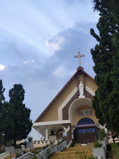 This image depicts the Domaine de Marie Church, also known as Mai Anh Church, located at 01 Ngô Quyền Street, Phường 6, Thành phố Đà Lạt, Lâm Đồng, Vietnam.
The scene presents an upward view of the church's facade under a partially cloudy, light blue sky. The church features a steep, reddish-brown tiled roof culminating in a white cross at its apex. Below the cross, a white statue of a female figure, presumably Mary, stands within a recessed niche. The main facade is primarily painted a light beige/tan with brown and white architectural trim. A large arched entrance dominates the lower central part of the facade. Above the arch, a colorful mosaic panel depicts a religious scene, likely "The Last Supper." Below the mosaic and above the dark wooden double doors, a banner with Vietnamese text reads "TÔI SẼ TRỞI DẬY VỀ CÙNG CHA TÔI." To the left of the main entrance arch, a white relief sculpture of an angel with outstretched wings is affixed to the wall. Further left, a covered porch with white pillars extends from the building.
A wide, multi-tiered staircase, appearing golden-yellow from discolored steps, leads directly up to the church entrance. White railings with grey posts line both sides of the staircase. Green foliage and shrubs are planted alongside the stairs and on adjacent terraced areas. Tall, dark green coniferous trees, resembling cypress or pine, frame the church on both the left and right sides, partially obscuring the upper portions of the sky. No individuals are visible within the frame.