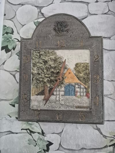This is a close-up, eye-level, vertical shot of an ornate, decorative clock mounted on a wall with a stone-patterned wallpaper. The clock itself is shaped like a stylized house with a rounded left side. The frame of the clock is a dark, textured metal, possibly bronze or iron, with embossed Roman numerals marking the hours from 1 to 12 around its perimeter.
The face of the clock depicts a miniature, three-dimensional scene. A rustic house facade with a blue door is visible, surrounded by textured greenery representing bushes and trees. A small, conical, straw-colored roof element protrudes from the house. The clock hands are made of a reddish-brown material and are shaped like stylized wooden objects, one resembling a propeller. The hour hand is pointing roughly to the 10 o'clock position, and the minute hand is pointing to the 1 o'clock position, indicating a time of approximately 10:05. There is a small cluster of dark red berries or flowers on the house. On the left side of the clock's frame, a textured embellishment resembling a sprig of lavender is present.
The wallpaper behind the clock has a pattern of irregular, light-colored stones, giving the impression of an exterior wall. Some green vine-like foliage with light green leaves is visible in the lower and right portions of the frame, partially obscuring the wallpaper. The lighting appears to be from an indoor source, casting a soft, even light on the scene, with no strong shadows. The overall mood is quaint and decorative, with a hint of rustic charm.
There is some visible text on the clock: "JUN CHANS" is embossed vertically on the right side of the clock face's frame. There are also small numbers, possibly a serial number, "0004G" faintly visible near the bottom right of the clock face. The location context provided is Trier, Germany.