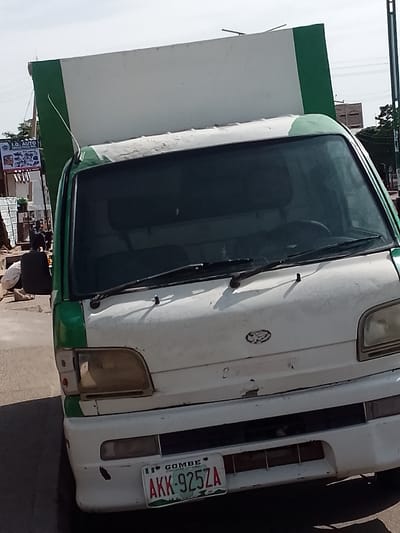 An up-close, eye-level, medium shot captures a white and green delivery truck parked on a paved street, likely in Pantame, Nigeria. The truck occupies the majority of the frame, with its front grille, headlights, windshield, and the side of its cargo bay visible. The vehicle appears weathered, with some rust and peeling paint. A license plate on the lower front of the truck reads "GOMBE AKK-925ZA". In the background, partially obscured by the truck, is a glimpse of a billboard advertising "I.G. AUTO", and a person, dressed in a black top and light-colored trousers, is crouched down on the left side of the image. The sky above is bright and clear, suggesting daytime. The overall impression is of a busy street scene in a Nigerian city.