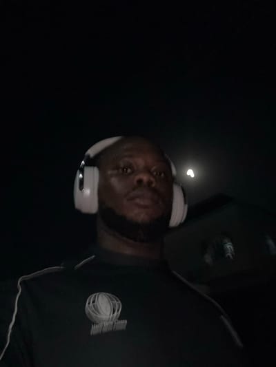 The image shows a man with dark skin and a beard wearing white headphones. He is facing the camera and slightly tilted. He is wearing a black shirt with a white logo on the chest. The background is dark, with a small crescent-shaped moon visible in the upper left. It is not possible to verify the location in the city of Lugbe, Nigeria from the image.