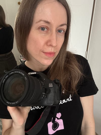 An indoor image depicts a female individual in Kudrovo, Russia, captured in a selfie. The woman, appearing to be in her 30s or 40s, has fair skin, light brown hair, and blue eyes, with a light pink tint on her lips. She wears a black short-sleeved t-shirt featuring white text, partially reading "heart," and a pink camera icon on the chest. Her left hand holds a black Canon EOS 600D DSLR camera, equipped with a Sigma Zoom 17-50mm 1:2.8 EX DC OS HSM lens, directed towards the viewer. A black camera strap with a red stripe is visible. The woman's gaze is directed towards the point of capture. In the background, a large reflective surface on the left side of the frame displays a partial reflection of the woman's back, clad in a black top and dark-colored pants. The immediate environment consists of plain white walls, a small circular fixture on the upper left wall, and a vertical door frame or wardrobe edge on the right.