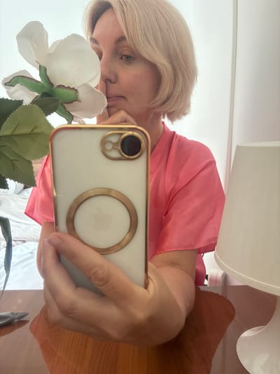 The image is a selfie taken by a fair-skinned woman with short blonde hair, wearing a bright pink collared garment. She is holding a white smartphone with a gold-rimmed case in her right hand, which is partially obscuring her face. The phone has a gold ring on its back. A white artificial flower with green leaves is held in front of her face, obscuring her left eye. She is looking towards the camera, with her right hand resting on her chin. The background shows a bed with white linens, a lamp with a white shade on a wooden surface, and a white wall. The lighting suggests an indoor environment, possibly a bedroom. There is no visible indication of the location being Gelendzhik, Russia.