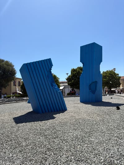 The image shows two abstract blue sculptures made of horizontal slats, positioned on a gravel-covered ground. The sculptures are large and appear to be hollow with cutouts, suggesting an artistic installation. One sculpture is in the foreground, angled towards the viewer, while the other is slightly behind and to the side.
In the background, there are trees and buildings, hinting at an outdoor urban setting. Some people are visible in the distance, gathered in front of a building, suggesting a public space or an event. The sky is clear and blue, indicating fair weather and likely daytime, possibly mid-morning or afternoon given the bright sunlight casting shadows.
The overall scene conveys a sense of modern art displayed in a public area. The blue color of the sculptures contrasts with the grey gravel and the natural green of the trees. The orientation of the photograph is landscape, and the perspective is slightly from above, looking down at the sculptures and the surrounding area. There is no visible text in the image. The location context of Florence, Italy, suggests this might be a contemporary art exhibit within the city.