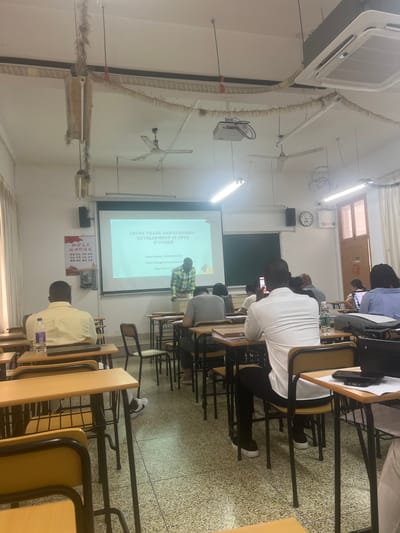 The image shows a lecture hall with students seated at desks facing a projector screen. The screen displays text related to "COCOA TRADE AND ECONOMIC DEVELOPMENT IN COTE D'IVOIRE". A presenter, a Black male wearing a checkered shirt, stands near the screen. Several students, predominantly Black males, are visible in the foreground and middle ground. One student in the foreground is using a mobile phone. The room features desks, chairs, fluorescent lighting, ceiling fans, and an air conditioning unit. There are also Chinese characters on a poster on the wall. The floor is tiled with a speckled pattern. The overall scene depicts an academic setting, likely a university lecture or presentation.