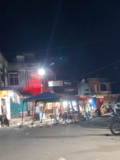 The scene depicts a street in Keudegeudong, Indonesia, at night. Street vendors are set up along the roadside. The street is littered with trash. A building with a red facade is visible in the background. The sky is dark, and power lines are visible. A motorcycle is parked nearby. People are present near the stalls and under the light fixtures. Banners are placed along the stalls.