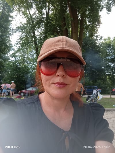 The image is a close-up selfie of a woman wearing a tan corduroy baseball cap and large red-tinted sunglasses. She has auburn hair and is wearing a black textured top. In the background, trees and a slightly overcast sky are visible, suggesting an outdoor setting. Behind her, there are several parked cars, some people in casual clothing, a stroller, and what appears to be a barbecue with smoke rising. The ground in the foreground is sandy. The date stamp on the image indicates June 20, 2026, at 5:02 PM. There is no information in the image to suggest the location is Wrocław, Poland.