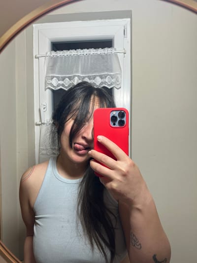 The scene is depicted as occurring within the city of Wüstenrot, Germany. The image is a mirror selfie showing a female individual from the chest up. The individual has dark hair that partially covers her face. Her left eye is visible, and her tongue is extended from her mouth. She is wearing a light blue ribbed tank top. A faint, multicolored linear mark is visible on her left shoulder. Her right hand holds a red smartphone, which obscures the right side of her face. Scars are present on the dorsal aspect of her right hand and distal forearm. A dark tattoo is partially visible on her right forearm. In the background, a white-framed window with a white, translucent lace curtain is visible behind her head. The wall to the right of the window is a plain, light color. A light-colored mirror frame is partially visible along the left edge of the image.