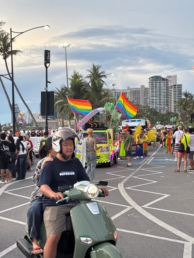 This is a daytime, eye-level shot of a Pride parade in Pattaya City, Thailand. The foreground features two individuals on a green scooter. The driver, wearing a dark blue t-shirt that reads "SUPERIOR" and a grey helmet, is in focus. Behind him, a person wearing a grey and blue plaid shirt is visible. The background shows a colorful parade with a lime-green float adorned with rainbow lights and decorations. Several individuals in elaborate costumes, including one with large yellow wings, are participating in the parade. Rainbow flags are prominently displayed, including one on the float and another in the distance. Other parade participants and spectators line the street, with tall buildings and palm trees visible in the background. The road surface has white markings, suggesting an urban environment. The sky is partly cloudy.