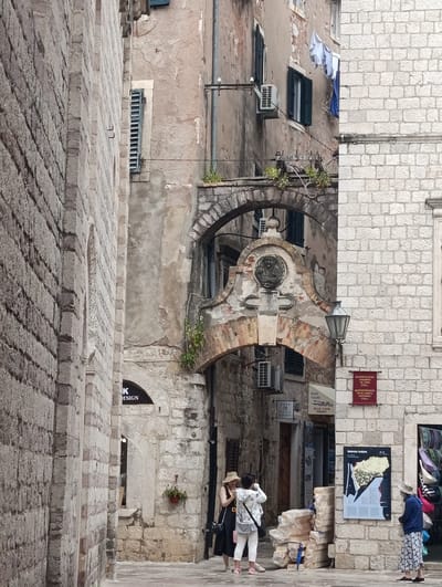 This image captures a narrow street scene in Kotor, Montenegro, likely during daylight hours under overcast skies. The setting is characterized by ancient stone architecture, with weathered buildings showcasing layers of history. The building on the left features rough-hewn, light-colored stone blocks, while the structure on the right is built with more uniform, lighter stone.

The focal point is an arched passageway that leads further into the street. Above this arch is a circular relief or medallion, and below it, a plaque with some text is visible. On the facade of the building to the right of the arch, a red sign with white lettering and a smaller white sign above it are affixed to the stone wall. Another sign, "BOAT TOURS," is visible to the left of the arch, along with "BLUE CAVE PERAST." Further left, a sign reads "K DESIGN."

Several people are present in the scene. Two women stand near the center of the image, one wearing a straw hat and a black dress, the other in a white jacket and white pants, appearing to take a photo. To the right, another person, wearing a patterned skirt and a blue top, stands looking at a large map poster mounted on the wall. In front of this poster, stacks of what appear to be building materials are piled up. A red flowerpot hangs on the wall to the left of the archway.

The overall impression is one of an old European town, with a mix of tourist-oriented businesses and historic architecture. The presence of air conditioning units on the buildings suggests a modern adaptation to the old structures. The clothing of the people and the general atmosphere hint at a warm day.