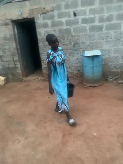 The media file shows a person standing in an outdoor area with a brick wall in the background and a dirt or earthen ground. The person is wearing a blue dress with a floral pattern. They are also wearing sandals with white stripes. The person's left leg is bent at the knee, and their right leg is extended. In the background, there is a large blue barrel with a white lid. To the left of the barrel, there is a dark opening, possibly a doorway or window, set into a rougher wall structure. The lighting suggests it is daytime, possibly late afternoon or early morning given the shadows. The overall scene appears to be in a modest, possibly rural or semi-urban setting. There is no visible text in the image.