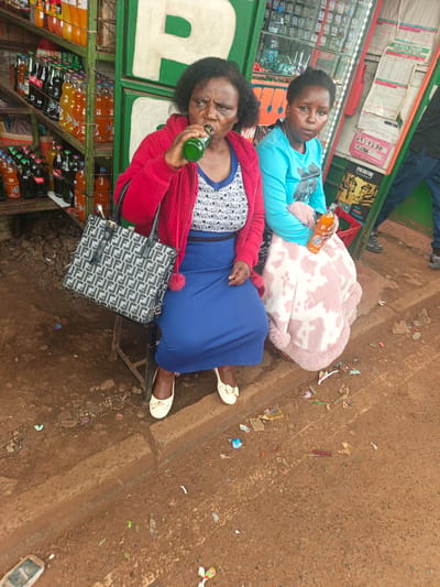 The image depicts two women seated outside a small street-side shop or market stall in Thika Township ward, Kenya. The older woman on the left, with dark skin and curly black hair, wears a red cardigan over a white patterned top, a blue skirt, and light-colored flat shoes. She is drinking from a green glass bottle and has a grey and black patterned tote bag, reminiscent of a Fendi design, by her side. To her right, a younger woman with dark skin and short black hair is dressed in a light blue long-sleeved top and wrapped in a pink and white patterned blanket. She holds an orange bottled drink. Both women appear to be resting.
Behind them, the shop structure is visible, painted green and white, with shelves fully stocked with various bottled beverages, including sodas like Coca-Cola and Fanta. To the right of the women, a display case and several signs are present. Visible text on these signs includes "M-PESA TARIFF EFFECTIVE 20TH JULY 2015," "AGENT CODE 6919230 LYDIA," and an advertisement for "PREDATOR NEW MANGO FLAVOUR." The ground is unpaved, dusty, and reddish-brown, with a concrete curb separating the sitting area from the path, and scattered litter. The scene takes place during the daytime, under natural light. The older woman has a focused expression, while the younger woman looks towards the camera with a neutral gaze. A partial view of a person in blue jeans is visible on the far right.