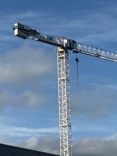 An image shows a construction crane against a blue sky with scattered clouds. The crane is white and has the word "walls" visible on its jib. The jib extends from the left side of the image towards the right. A dark, sloped roofline is visible in the bottom left corner of the image. The image does not contain any information about a specific location in the city of Cork, Ireland.