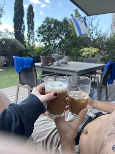 The image depicts an outdoor residential patio and garden scene in Ramat HaSharon, Israel, under a daytime sky with scattered clouds. Two individuals are partially visible from a low angle, holding clear glasses of iced coffee. The person on the left presents a hand with dark red manicured nails and a silver ring on the middle finger, wearing a dark long-sleeved garment and light-colored shorts. This hand holds a glass containing iced coffee topped with a thick layer of white foam. The person on the right displays a bare, hairy abdomen and a hand, wearing light-colored shorts with a visible dark belt. This hand holds a glass of iced coffee with visible ice cubes and less foam. The glasses are held together, indicating a toast or clinking action.
In the background, a light-colored rectangular outdoor dining table is visible, adorned with several potted plants and a blue and white decorative ceramic bowl. A blue towel or piece of clothing is draped over a dark-colored chair situated to the left of the table. Beyond the patio, a lush green garden extends with various trees and shrubs, including two tall, slender cypress-like trees on the left. An Israeli flag, featuring blue stripes and a Star of David on a white background, is partially visible, unfurled against the green foliage in the upper right portion of the frame. A grey awning or roof structure is visible at the very top. The ground beneath the furniture consists of both green lawn and a section of light-colored wooden decking.