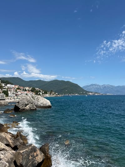 The image shows a coastal view of Herceg Novi, Montenegro. In the foreground, waves crash against large, gray rocks. The sea is a deep blue, with white foam from the waves. In the mid-ground, a large, rough-textured gray rock juts out of the water. Behind it, a cluster of white buildings with red-tiled roofs cascades down a hillside, forming the town of Herceg Novi. Lush green mountains rise behind the town, their slopes covered in vegetation. The sky is a bright blue with a few scattered white clouds. In the distance, a range of lighter, rocky mountains can be seen. Several small boats are dotted on the water. The scene depicts a sunny day with clear weather, showcasing the natural beauty and urban development of the coastal city.