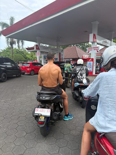 The image depicts a crowded Pertamina gas station in Munggu, Indonesia, under an overcast sky. In the foreground, a shirtless male with a muscular back is seated on a black Yamaha NMAX scooter (license plate DK 6382 XX), facing away from the camera. He wears black shorts and blue and green athletic shoes. To his right, another individual wearing a light blue, long-sleeved shirt is partially visible, seated on a red scooter. Behind the shirtless man, a person wearing a dark grey t-shirt with a "Nirvana" graphic and a white helmet is seated on another black scooter, also facing away.
Vehicles are queued for service. To the left, a black SUV is parked, and a red hatchback car is positioned further back. The gas station structure features a red roof supported by white pillars, with multiple fuel pumps visible. Signs identify fuel types such as "Pertamax," "Pertamax Dex," and "Pertalite 92." A prominent sign in the center indicates "JALUR KHUSUS PERTAMAX PERTAMAX" with directional arrows. The ground is paved with hexagonal grey bricks. Palm trees and dense green foliage are visible behind the station, along with traditional-style buildings featuring terracotta-tiled roofs.