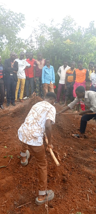 This outdoor image depicts a group of men gathered in a rural area of Fizi, Democratic Republic of the Congo. The scene is dominated by reddish-brown soil, suggesting rich earth, with dense green vegetation and trees visible in the background under an overcast or bright sky.
In the foreground, two men are actively engaged in digging or moving soil. The man on the left, facing away from the camera, wears a white patterned shirt, brown trousers, and grey sandals, and is using a long wooden stick-like tool to manipulate the earth. To his right, another man, wearing a light-colored long-sleeved shirt and dark trousers, is also using a similar wooden tool, appearing to level or clear the ground. A pile of loose soil is prominent between them.
Behind these two individuals, a larger group of approximately ten to twelve men stands observing the activity. They are dressed in a variety of casual attire, including t-shirts, button-up shirts, and trousers. One man in the background wears a white t-shirt with the text "Emirates Fly Better" clearly visible. Another man in the observing group wears a dark suit jacket and glasses. Their expressions appear attentive and serious.
The overall atmosphere suggests manual labor or community work taking place during the daytime under cloudy conditions. The ground is uneven, indicative of recent digging or groundwork.