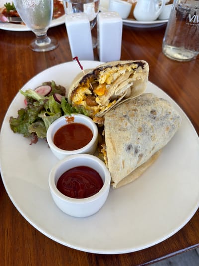 The image captures a meal served on a dark reddish-brown wooden table at a restaurant in San Pedro Town, Belize. A white oval plate dominates the central foreground, presenting two burritos. One burrito is cut in half, revealing a filling of scrambled eggs, golden-brown pieces (possibly sausage or potatoes), and a dark, likely bean-based, spread. A red plastic toothpick is inserted into the cut burrito. The second burrito is wrapped whole. Alongside the burritos, a small side salad features green leafy lettuce and sliced red radishes. Two small white ramekins are present on the plate; one contains a reddish-brown sauce, possibly salsa or hot sauce, while the other holds bright red ketchup. In the blurred background, a clear drinking glass with condensation, two white rectangular shakers (likely salt and pepper), and a clear bottle labeled "VIVA ADVANCED" are visible. A partial view of another white plate with food and a human hand is discernible in the upper left background, suggesting a companion diner. Other white ceramic dishes are faintly visible in the far background.