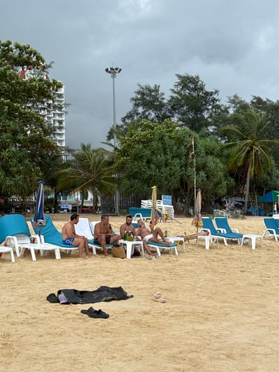 A group of people are relaxing on a beach in Karon, Thailand. The scene is set on a sandy beach with rows of white plastic lounge chairs and blue fabric. Several people are seated in the chairs, some with drinks. Palm trees and other lush green vegetation are visible behind the chairs, and a tall building complex is seen in the distance. The sky is overcast and cloudy, suggesting a possibility of rain or a storm. A street lamp or light tower stands tall in the background. Some clothing items, including a black garment and a pair of black sandals, are lying on the sand. The overall mood appears relaxed, with individuals enjoying their time at the beach despite the cloudy weather.