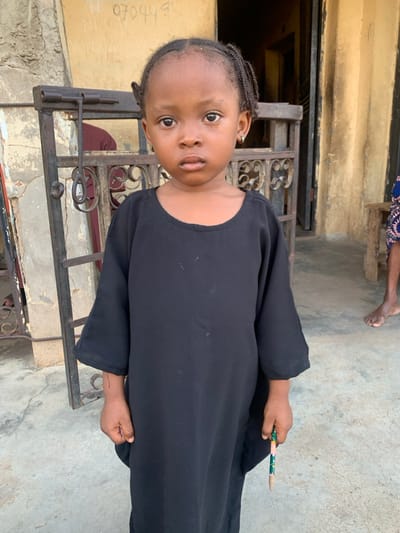 The image depicts a young child standing outdoors in Okene, Nigeria. The child, with dark skin, dark braided hair, and light-colored stud earrings, faces the camera directly with a neutral expression. They wear a loose-fitting, long-sleeved black garment. In their left hand, the child holds a short, green and pink patterned pencil with a sharpened tip.
The background features a building with weathered, light-colored walls. To the viewer's left, a dark metal security gate or grille is partially visible, behind which a person in dark red clothing is partially obscured. Faint dark script, including "070449", is visible on the wall above the child's head. To the viewer's right, a dark doorway is present, and a portion of another person's bare legs and feet, seated on a wooden bench, are visible against an ochre-colored section of the wall. The child stands on a light grey, paved surface.