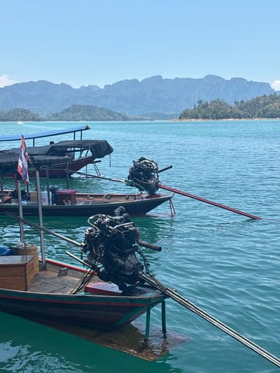 The image shows two traditional Thai longtail boats docked in Cheow Lan Lake, located in Ban Ta Khun, Surat Thani Province, Thailand. The boats are equipped with large outboard motors mounted on long poles, which are typical of this type of vessel. In the background, there are limestone karst mountains. The water is turquoise. The sky is pale blue.