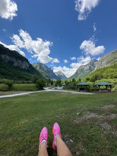 A person's legs, wearing bright pink sneakers and white socks, are visible in the foreground, resting on a grassy area. The background opens up to a scenic view of a valley with lush green mountains on either side. A clear blue sky with scattered white clouds stretches above. In the mid-ground, there's a gravel path leading towards a serene body of water, possibly a river or lake. Two rustic wooden gazebos with green roofs are situated near the water, offering a place for rest. The overall environment suggests a natural park or recreational area in Vusanje, Montenegro, characterized by its mountainous landscape and clear weather.