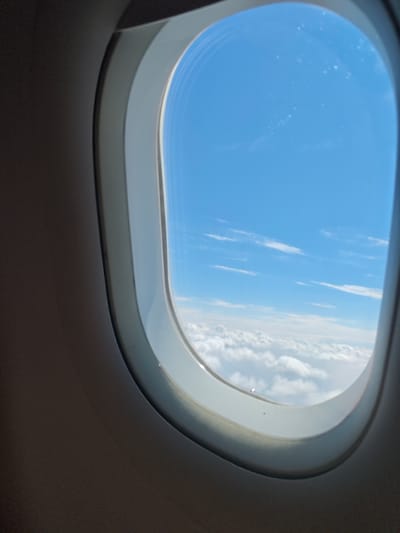 An airplane window frames a view of a bright blue sky dotted with wispy white clouds. Below the clouds, a dense layer of fluffy white clouds stretches out, obscuring any view of the ground. The interior of the airplane cabin is visible around the window, with dark gray paneling and the white frame of the window itself. The scene appears to be an aerial view from a flight.