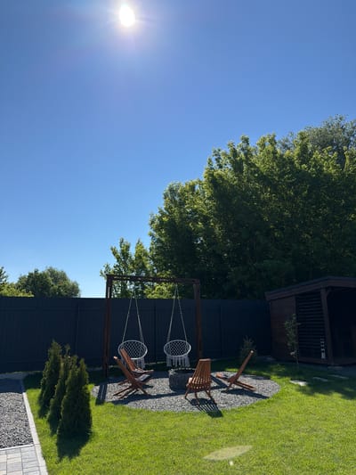 The image depicts a backyard in Sharovechka, Ukraine, on a bright, sunny day with a clear blue sky. The scene features a fire pit area surrounded by two wooden Adirondack chairs and a gravel base. Above the fire pit, two white macrame swings hang from a wooden frame. To the left, a row of three conical evergreen trees stands on a well-maintained lawn, bordered by a gravel path. A dark wooden fence encloses the property. To the right, a wooden gazebo structure with a dark roof is visible. The overall impression is one of a peaceful, outdoor recreational space.