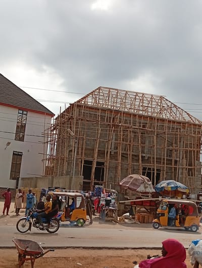 The image depicts a street scene in Masaka, Nigeria. An unfinished building with extensive bamboo scaffolding dominates the background. To the left stands a completed two-story building with windows. In the foreground, a street is visible with several vehicles: a motorcycle with two riders, and two yellow auto-rickshaws (tuk-tuks). People are standing along the roadside, and a market stall is set up with goods displayed under umbrellas. The weather appears overcast. A wheelbarrow sits in the foreground to the left.