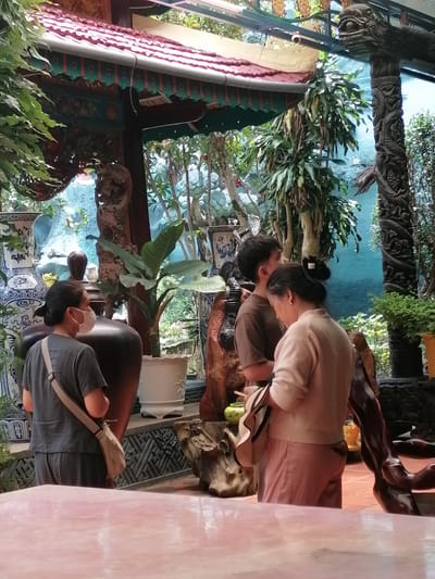 The image depicts an outdoor scene in Xuân Thành, Vietnam, possibly a garden or courtyard area, characterized by lush vegetation and traditional decorative elements. Three individuals are present. In the foreground, a person wearing a grey t-shirt and grey pants, with a light-colored cross-body bag, stands with their back to the camera, facing towards the right. They are wearing a light-colored face mask. To their right and slightly behind, a young person wearing a brown t-shirt is partially visible, facing away from the camera. Next to them, a person wearing a light pink sweater and pink pants is facing their left, looking down. They are holding a straw hat in their left hand.
The environment features ornate architectural elements, including a roof with red tiles and decorative green trim. A tall, intricately carved wooden pillar with a dragon motif stands prominently on the right side of the frame. Several large ceramic vases, one with a blue and white pattern, are visible. Potted plants and large planters are scattered throughout the scene, contributing to a dense, verdant atmosphere. The ground appears to be tiled or paved with reddish material. A pinkish-red reflective surface, possibly a table, occupies the bottom portion of the image. The overall impression is of a culturally rich and visually detailed outdoor space.