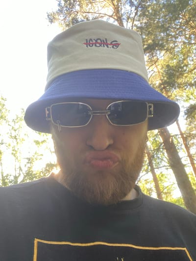 The media file is a close-up, low-angle selfie of a person wearing a bucket hat and sunglasses outdoors. The subject has light-colored hair and a beard. The bucket hat is light-colored with a blue band and features red embroidered text. The sunglasses are rectangular with metal frames. The person is looking upwards and appears to be in a relaxed or playful mood.

The setting is outdoors, with sunlight filtering through trees, suggesting it is daytime. The foliage indicates a natural environment, possibly a park or a wooded area. The sky is bright and washed out due to the sun's position. The person is wearing a dark crewneck sweatshirt with a yellow embroidered detail on the shoulder.

The text visible on the bucket hat appears to be "SNOOP" or a similar stylized word. The overall impression is of a casual outdoor activity on a sunny day. The specific location cues within Zheleznogorsk, Russia, are not explicitly discernible from the image itself beyond the general outdoor, wooded setting.
