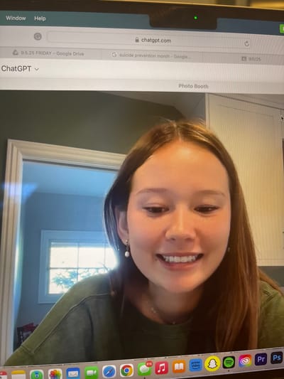 This image depicts a laptop screen displaying a young female in a video feed, likely from the Photo Booth application, centered within the frame. The scene is described as being in Rolling Hills Estates, United States.

The female, appearing to be in her late teens or early twenties, has long, straight, dark brown hair parted down the middle. She is smiling broadly, revealing upper and lower teeth, and wears a solid olive-green long-sleeved top and small, dangling pearl earrings. A delicate necklace is also visible.

Above the video feed, web browser tabs are open, indicating a Google Chrome interface. The active tab displays "chatgpt.com" in the URL bar and "ChatGPT" as the tab title. Other visible tabs include "9.5.25 FRIDAY - Google Drive" and "suicide prevention month - Google...". A small green light above the screen confirms the laptop's camera is active.

The background visible in the video feed shows an indoor environment. To the viewer's left, a doorway with white trim leads to another room, where a bright window reveals outdoor foliage. To the viewer's right, a white cabinet or wall section is visible. The wall behind the female appears to be a muted green color.

At the bottom of the laptop screen, a macOS dock displays various application icons, including Safari, Chrome, Messages (showing 816 unread notifications), Music, Books, Prime Video, Snapchat, Spotify, and Photoshop.