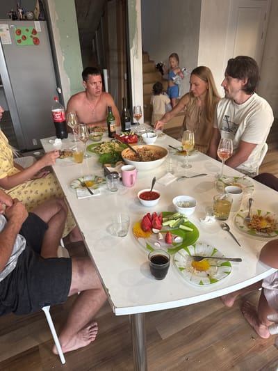 The photograph captures a gathering of people seated around a white table, likely in a dining area or a communal space. The setting appears to be an interior with a refrigerator visible on the left, adorned with decorative magnets. The overall ambiance suggests a meal or social event in progress.
At the table, six individuals are partially visible, engaged in eating and interacting. A man, shirtless and wearing a necklace, is seated on the left side of the frame, looking to the side. Next to him, a woman with blonde hair, wearing a beige dress, is leaning forward, appearing to interact with the food on the table. To her right, a man with dark hair, wearing a t-shirt, is looking towards the woman. In the background, on a staircase, two young children are visible, one in blue shorts and the other in a beige outfit.
The table is laden with food and drinks. Various dishes, including salads, rice dishes, and bowls of what appears to be sauce or dip, are spread across the surface. Several glasses, some containing drinks, are also present. A bottle of Coca-Cola and two bottles of wine are among the beverages. Plates with remnants of food and used cutlery indicate that the meal is underway or has recently concluded. The plates themselves are decorated with a floral pattern.
The flooring is made of wooden planks, and the walls exhibit unfinished construction elements, suggesting the location might be undergoing renovation or is a rustic dwelling. The lighting is ambient, with no single dominant light source. The image is taken from a slightly elevated angle, looking down at the table. The overall scene conveys a casual, familial gathering. The location is identified as Verkhniy Yurt, Russia.