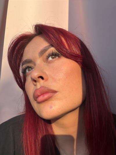 A close-up, low-angle portrait shows a woman with dark red hair and fair skin with freckles looking upwards. She is wearing a black top and has prominent eyelashes. The lighting suggests it is either sunrise or sunset, casting a warm glow on her face and creating a shadow line across her right cheek and neck. The background is a wall with a soft pink to purple gradient. The image does not contain any recognizable landmarks or elements that would indicate a specific location within the city of Uzukovo, Russia.