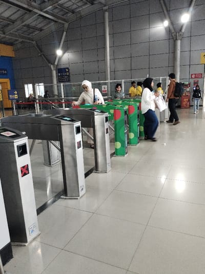 The image shows a group of people walking through a series of turnstiles at a train station in Bekasi Regency, Indonesia. The coordinates of the station are -6.259 latitude and 107.056 longitude. There are seven turnstiles in total, with each one having a green and red sticker on it.  The turnstiles are made of metal and appear to be fairly new. The person closest to the camera is a woman wearing a white headscarf.  She is holding a blue bottle in her left hand and is using her right hand to swipe a card on the turnstile. Behind her, there is a man in a yellow shirt and a woman in a blue shirt.  The floor is tiled in a light grey color. The walls are gray and appear to be made of concrete blocks. The lighting in the room is bright.  The image appears to be taken from a slightly elevated position. There is a red line on the floor that runs parallel to the turnstiles.  A second line is visible in the background which leads to a set of escalators. The image is focused on the people walking through the turnstiles.