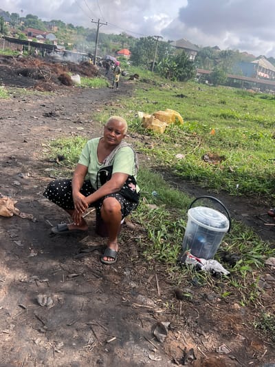 In an outdoor environment in Obosi, Nigeria, a woman is crouched on the ground, appearing to examine her feet. She wears a light green top, a black skirt with white stars, and gray sandals. A transparent plastic bucket is located to her right. The ground appears muddy and soiled, with remnants of burning debris. In the background, there are houses, a power pole, and other people. There is smoke in the distance, suggesting a recent fire. The sky is overcast.