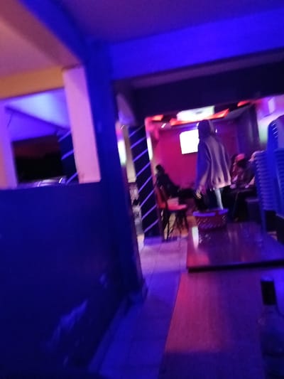 A dimly lit interior space, possibly a bar or lounge, features blue and purple ambient lighting. A counter runs along the right side, with a partial view of a bottle. Several people are seated in the background, some at tables with chairs, near a glowing screen or sign. The décor includes dark pillars with diagonal stripes and a red-lit wall. The overall impression is that of a relaxed, intimate setting.