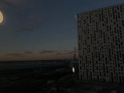 A nighttime, wide-angle shot captures an urban industrial landscape at dusk. The foreground is dominated by a large, modern, multi-story building with a grid-like facade of windows and dark vertical accents. To the left of the building, a tall industrial smokestack with red and white bands stands against a dusky sky filled with soft, pink-tinged clouds.

In the middle ground, a vast expanse of low-rise industrial buildings with flat, reflective roofs stretches across the scene. Several trucks and what appear to be shipping containers are visible on the grounds. A street runs through the lower part of the frame, with a few vehicles, including a white van and a red car, parked or driving.

The scene is dimly lit, suggesting either the very early morning or late evening. A bright, oval-shaped light fixture or reflection hovers in the air between the smokestack and the modern building, adding an ethereal quality to the image. The overall atmosphere is one of industrial activity under a fading sky. There is no visible text.