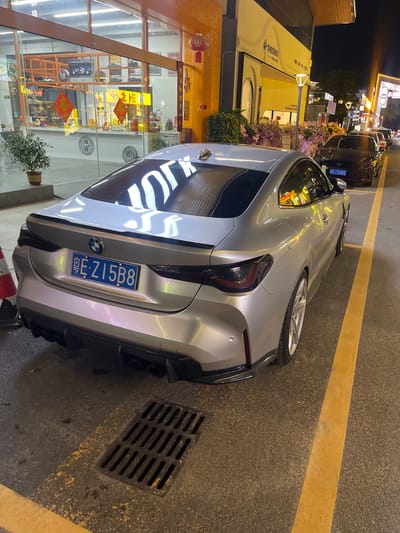 The image shows a silver BMW M4 parked on a street in Panyu District, China. The car is facing away from the camera, and its license plate is clearly visible. The license plate reads "E-Z15B8". The car is parked next to a yellow line on the asphalt. In the background, there is a building with a glass storefront, and several other cars are parked in a line. The scene appears to be at night, as the lights are on in the buildings and streetlights are visible. The overall environment is urban, with a commercial building and a street.