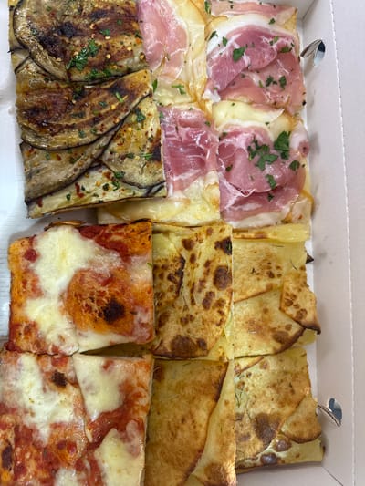 The image shows a box of different pizza slices. The top row features slices with prosciutto and parsley, potato slices, and potato slices with rosemary. The bottom row has eggplant slices, tomato and mozzarella slices, and slices with tomato and mozzarella. The pizzas are inside a white cardboard box. The location is Aprilia, Italy.