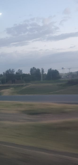 A cloudy sky dominates the upper-left portion of the scene. A highway runs vertically down the middle. Vegetation, possibly fields or grass, is blurred on the right side, indicating motion. Buildings, trees, and utility poles are visible in the distance on the left of the highway, set against the skyline. The blurring suggests the photograph was taken from a moving vehicle.
