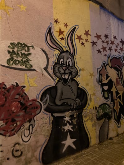 The image displays graffiti on a wall in Badalona, Spain. The graffiti features a cartoon rabbit character, resembling Bugs Bunny, depicted in a black top hat with white stars. To the left of the rabbit, there is green graffiti in a bubble speech that reads "SMART AUX COWS." To the right of the rabbit, there is more graffiti in various colors, including orange, yellow, and blue, with abstract shapes. Further to the right, a stylized mushroom with a green cap is painted on the wall. The wall itself is textured and shows signs of wear and tear, with peeling paint in several areas. The ground in front of the wall appears to be paved with brickwork. The lighting suggests it is nighttime or a dimly lit area, possibly an alleyway or underpass, with artificial light casting shadows and illuminating the graffiti. The graffiti appears to be on an exterior wall, possibly part of a building or urban infrastructure.