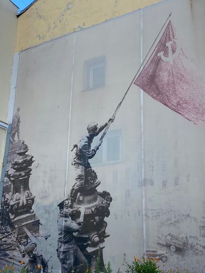 This image depicts a mural on the side of a building in Novokuybyshevsk, Russia. The mural is a grayscale representation of historical figures and a large red flag with a sickle and hammer symbol. The central figures are two soldiers, one in the foreground hoisting the flag atop a damaged monument, and another soldier to the left and slightly lower, also appearing to be involved in the scene. The background shows what appears to be a city under duress, with faint outlines of buildings and possibly rubble. A large red flag, rendered with a textured, chalk-like appearance, dominates the upper right portion of the mural, its pole held by the soldier. The building itself has a weathered, pale yellow facade with peeling paint at the top. Orange marigolds are blooming at the bottom of the mural.