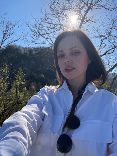 A selfie of a woman outdoors in the city of Bled, Slovenia. The woman has long brown hair, and is wearing a white button-down shirt. She has sunglasses hanging from the collar of her shirt. In the background, there are trees and the sky. The sun is shining brightly in the upper left corner of the frame, creating a lens flare.