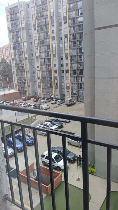 This high-angle, outdoor image captures a residential complex in Bogota, Colombia, under an overcast sky. The viewpoint is from a balcony, with a black metal railing comprising vertical bars traversing the lower foreground.
The midground features an expansive concrete parking lot containing numerous parked vehicles of various types and colors. Specific vehicles identifiable include a white Ford EcoSport SUV with "0 659" marked on its side, a dark grey sedan, two blue sedans, a silver pickup truck, a white box-body delivery truck, and a white van, possibly a service or transport vehicle. Yellow cylindrical bollards are sparsely distributed across the parking area. To the left of the parking lot, a half-basketball court with white markings and a hoop is visible next to a low, brown building. A rectangular brick structure, open at the top and featuring an internal staircase descending below ground level, occupies the lower-left quadrant of the midground. Patches of green artificial turf or grass are present adjacent to the brick structure and along the base of the buildings. One individual, wearing a dark top, blue jeans, and carrying a backpack, is observed walking on a concrete pathway towards the right-side building.
The background is dominated by multiple high-rise apartment buildings. The most prominent structure, centrally located, is a tall, multi-story building characterized by a light grey facade, distinct yellow vertical architectural accents, and numerous balconies with black railings. Several balconies display various personal items. A closer, grey concrete building with visible horizontal floor lines partially frames the right side of the image. In the far left distance, a shorter, reddish-brown apartment building is partially visible, interspersed with trees. The sky is uniformly overcast, indicating a cloudy daytime environment.